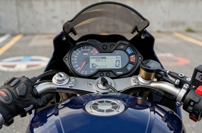 Gros plan sur le tableau de bord numérique et le guidon d'une Yamaha R6 avec des gants de moto en cuir posés dessus